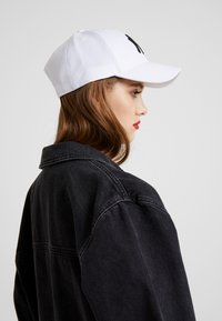 Hvid baseballkappe med sort logo, båret med en sort denimjakke. Har en struktureret form og synlige syninger.