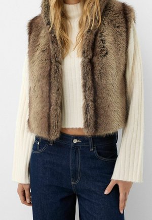 Femme portant un gilet sans manches en fausse fourrure marron par-dessus un pull court blanc côtelé à manches longues et un jean bleu taille haute.