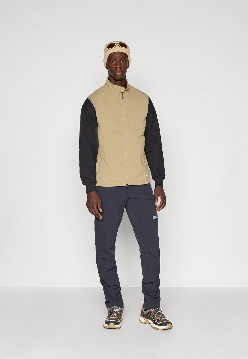 Giacca zip-up beige e nera con parte superiore testurizzata e maniche lisce, abbinata a pantaloni scuri e scarpe robuste con suola chiara.