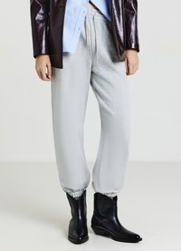 Persona che indossa pantaloni tuta grigi chiari, stivali di pelle nera, una camicia blu e una giacca lucida marrone scuro su uno sfondo semplice.