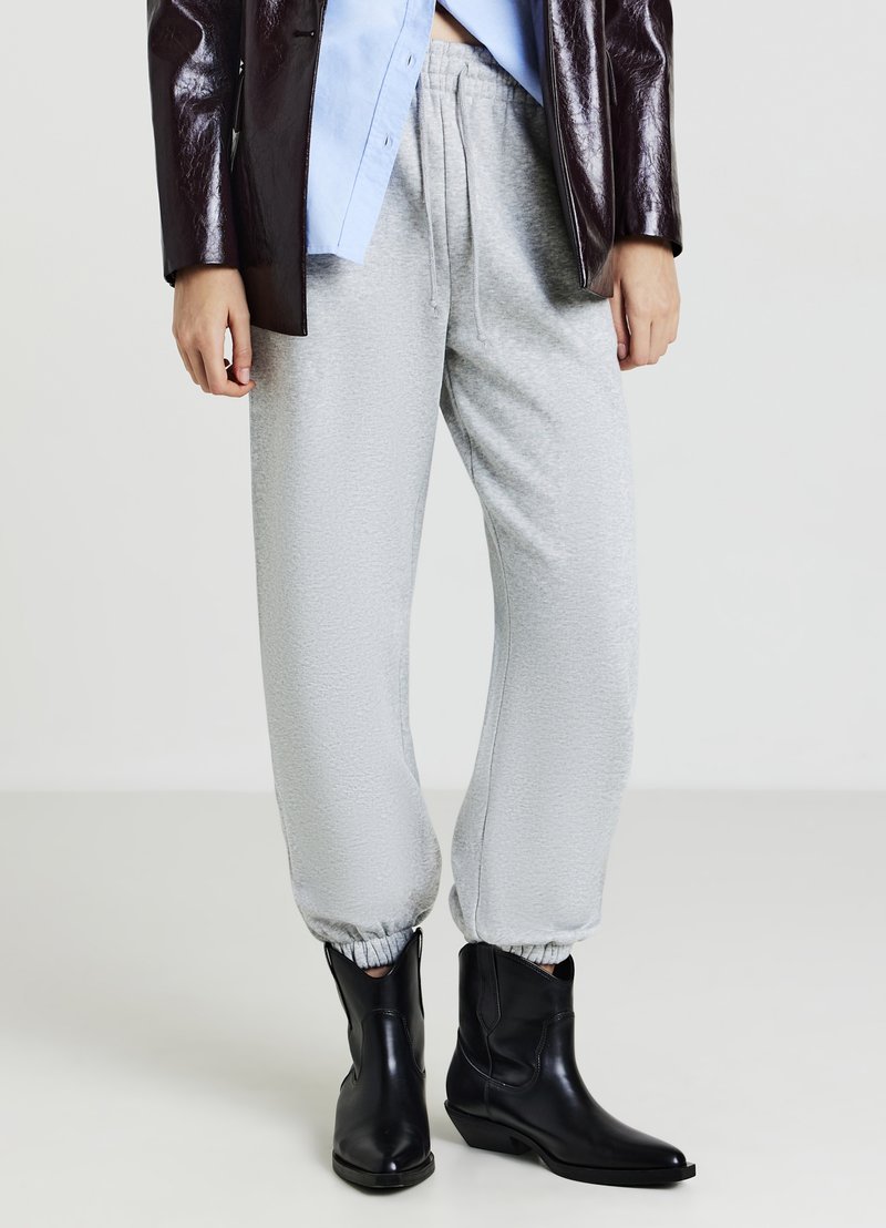 Persona che indossa pantaloni tuta grigi chiari, stivali di pelle nera, una camicia blu e una giacca lucida marrone scuro su uno sfondo semplice.