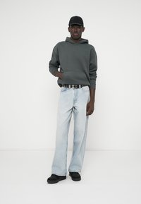 Szara bluza z kapturem, jasnoniebieskie dżinsy o luźnym kroju, czarny pasek z srebrnymi ozdobami i czarne buty. Model ma na sobie dopasowaną czapkę z daszkiem.