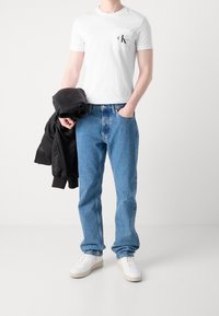 Muž nosící bílou tričko Calvin Klein, modré džíny, bílé tenisky, držící černou bundu v jedné ruce, stojící proti bílému pozadí.