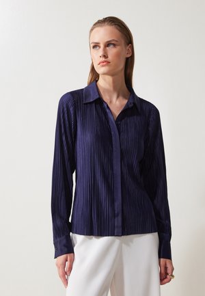 Camicia plissettata blu navy con colletto classico, chiusura frontale con bottoni e maniche lunghe. Texture liscia con dettagli a contrasto sui polsini.