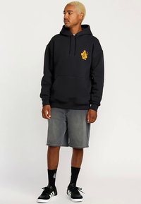 Svart huvtröja med framficka, gul grafisk design på vänster bröst, matchad med gråa jeansshorts och svarta sneakers med vita detaljer.