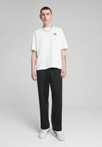 Hvid t-shirt med korte ærmer og et sort logo på venstre bryst, kombineret med sorte joggingbukser med en hvid side-stribe. Hvide sneakers.