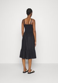 Robe noire sans manches avec corsage smocké et ourlet à volants. Le modèle est pieds nus, mettant en valeur une coupe décontractée et une texture tissée.