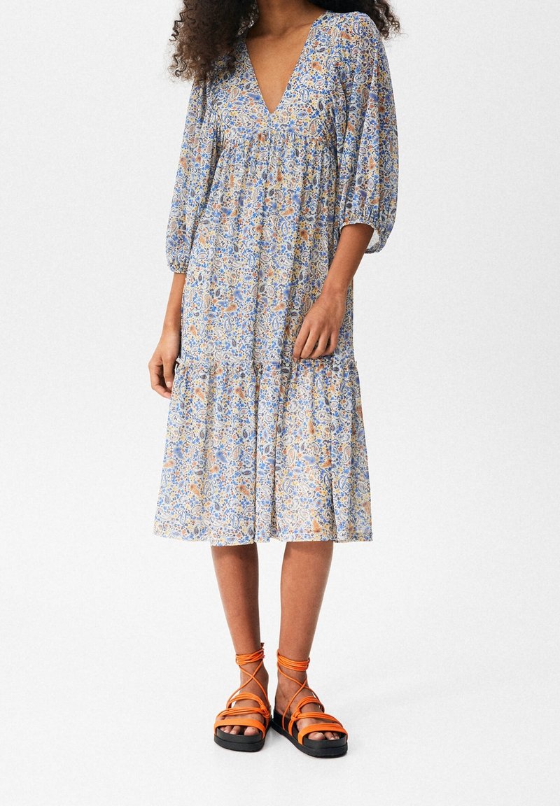 Femme portant une robe fleurie au genou avec manches trois-quarts et un profond décolleté en V, assortie à des sandales orange à lacets.