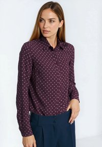 Bordowa koszula na długi rękaw z białymi kropkami, z kołnierzykiem i kieszenią po lewej stronie na piersi, w połączeniu z granatowymi spodniami.
