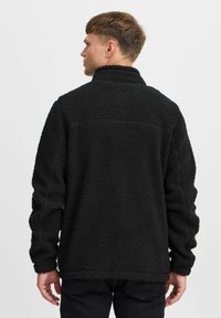Chaqueta de forro polar negra con un cuello alto, superficie texturizada y un corte relajado. El diseño incluye una costura en la parte superior de la espalda.