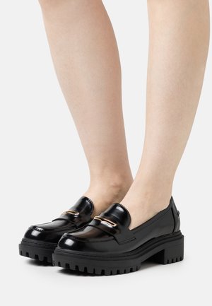 Black shiny leather loafers with chunky lug soles and gold metal accents worn on bare legs against a plain light background.