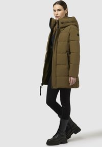 Oliven grøn pufferjakke med hætte, der har lynlåslukning, sidelommer og ribbede manchetter; båret over sorte leggings og kraftige sorte støvler.