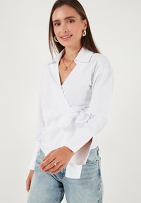 Young woman wearing a white wrap blouse tied at the waist, light blue jeans, gold hoop earrings, and a gold star necklace, smiling slightly.