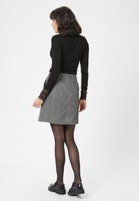 Vrouw die zijwaarts staat, gekleed in een zwart longsleeve top met kanten manchetten, een grijze herringbone rok, zwarte panty en zwarte loafers op een witte achtergrond.