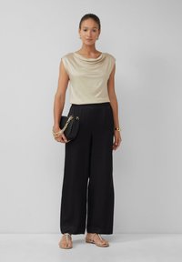 Beige drape top, zwarte wijde broek, gouden schoudertas met ketting, en zilveren slippers. Soepel stof en een ontspannen pasvorm.