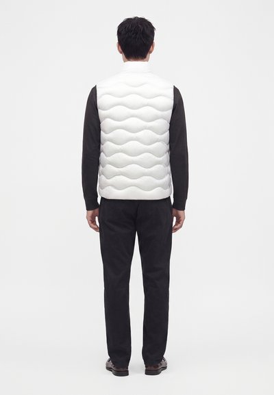 Hombre de pie con la espalda hacia el espectador, vistiendo un chaleco acolchado blanco, camisa negra de manga larga, pantalones negros y zapatos marrones.