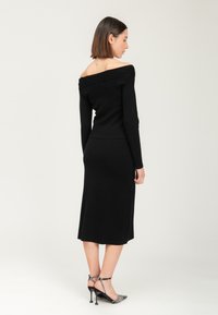 Abito nero a maglia off-the-shoulder con maniche lunghe, silhouette aderente e lunghezza al ginocchio; abbinato a tacchi alti a fantasia bianca e nera.