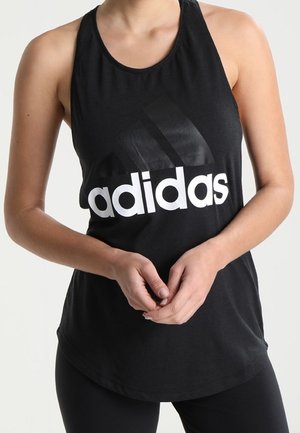 Kobieta ubrana w czarny, bezrękawnik Adidas z dużym białym logo, splecione ręce przed talią.