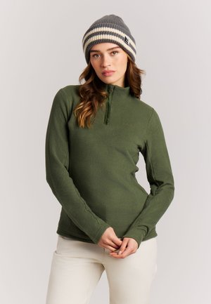 Femme portant un pull polaire vert et un bonnet rayé gris, debout, les mains jointes devant, sur un fond uni.