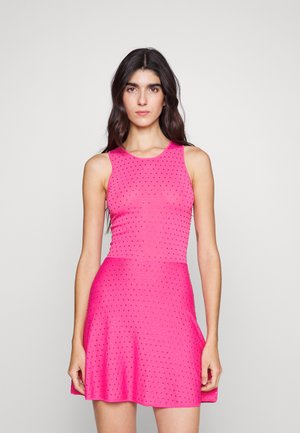 Nő áll szemben, ujjatlan, élénk rózsaszín ruhában, kicsi fekete pöttyökkel és bővülő szoknyával, egy egyszínű háttér előtt.