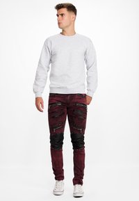 Sweatshirt gris avec des poignets côtelés, associé à un jean bordeaux déchiré avec plusieurs poches zippées et des accents en cuir noir. Baskets blanches.