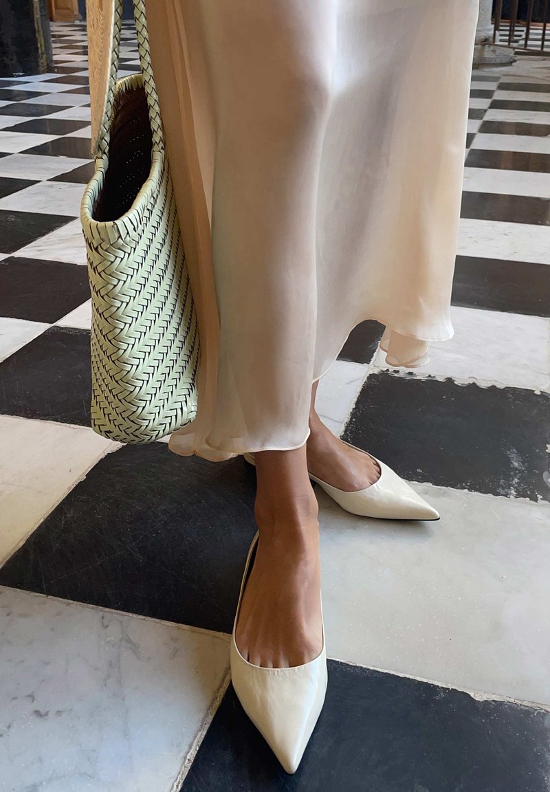 Personne portant des ballerines blanches pointues et une longue robe crème transparente, debout sur un sol à damier noir et blanc, tenant un sac tissé.