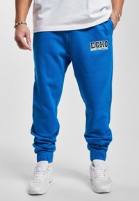 Pantaloni de trening albaștri cu talie elastică �și manșete. Prezintă un logo alb "ECKO" pe coapsa stângă. Materialul pare moale și texturat.