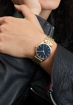 Montre en or avec un cadran noir et des marquages minimalistes, dotée d'un bracelet en maillons métalliques, portée sur un poignet au-dessus d'une veste en cuir noir.