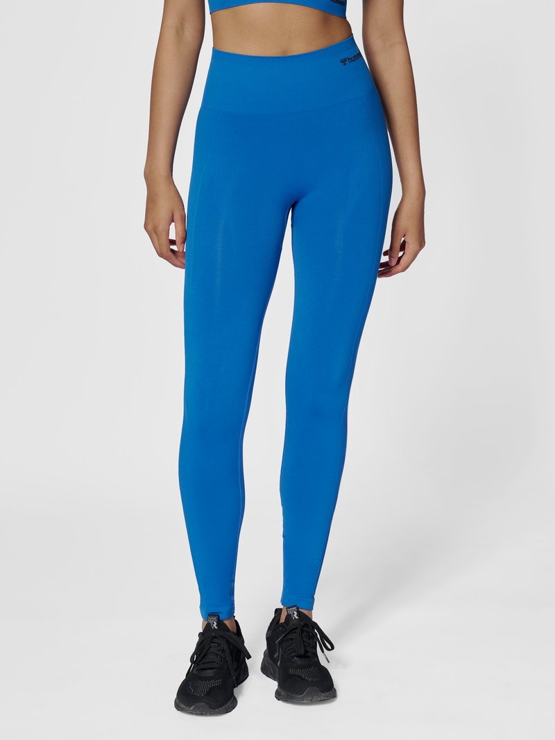 Vrouw draagt felblauwe high-waisted legging en zwarte sportschoenen, staand tegen een neutrale achtergrond.