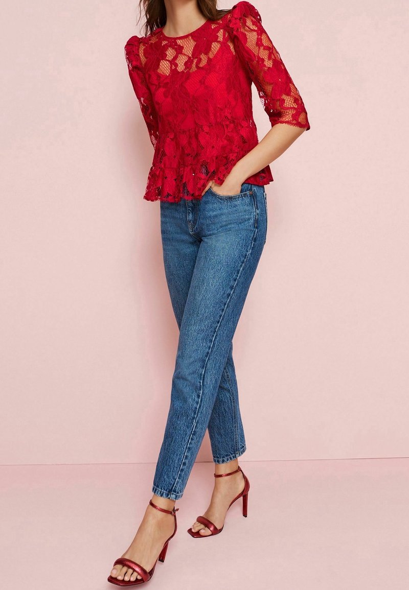 Haut péplum en dentelle rouge à manches trois-quarts, associé à un jean taille haute bleu clair et des escarpins rouges à bride à la cheville. Fond rose.