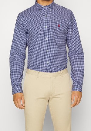 Homme portant une chemise à carreaux bleu marine et blanche boutonnée, rentrée dans un pantalon beige, debout devant un fond blanc uni.