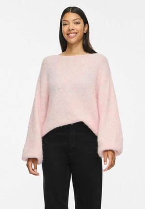 Jeune femme aux longs cheveux foncés portant un pull duveteux rose clair et un pantalon noir taille haute, souriante sur fond blanc.