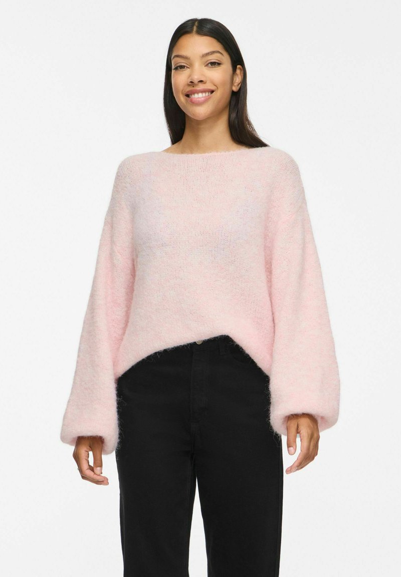 Jeune femme aux longs cheveux foncés portant un pull duveteux rose clair et un pantalon noir taille haute, souriante sur fond blanc.