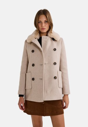 Manteau beige avec col en fausse fourrure, design croisé, grands boutons et poches avant. Porté par-dessus un haut noir et une jupe marron.