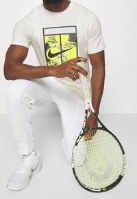 Camiseta gráfica de Nike en color blanco roto con un estampado amarillo y negro, combinada con pantalones deportivos blancos y una raqueta de tenis con un marco negro y cuerdas verdes.