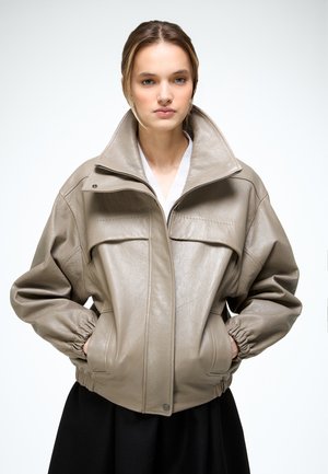 Junge Frau trägt eine beige Lederjacke mit hohem Kragen, die Hände in den Taschen, steht vor einem schlichten hellen Hintergrund.