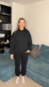 Moteris stovinti juodu gobtuvu ir kelnėmis su baltais sportbačiais gyvenamajame kambaryje šalia mėlyno sofos ir juodos lentynų sistemos.