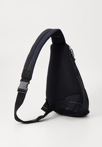 Svart axelremsväska med yta av texturerat nät och släta paneler. Utrustad med justerbar rem, dragkedjestängning och gröna sömnadsdetaljer.