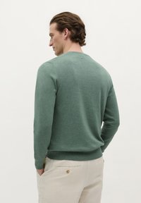 Groene gebreide trui met lange mouwen en een ronde halslijn, met een soepele textuur en een geribde zoom. G gedragen met lichtgekleurde broek.