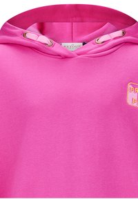 Roze capuchontrui van een gestructureerde stof, met metalen oogjes en een patch met "PROUD" in contrasterende kleuren.