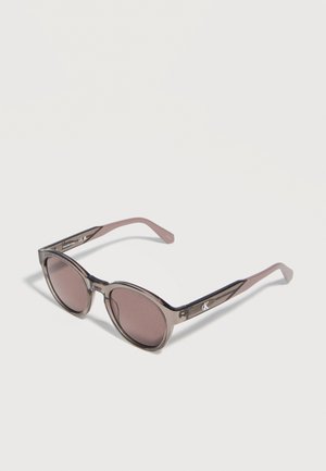 Apvalūs akiniai nuo saulės su permatomais pilkos spalvos rėmeliais ir rožinės spalvos filtro stiklais, ant šoninių rankenėlių matomas mažas „CK“ logotipas.