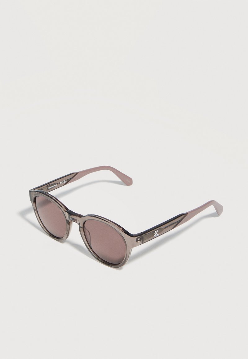 Στρογγυλά γυαλιά ηλίου με διαφανή γκρι πλαίσια και ροζ φακούς με απόχρωση, που φέρουν μικρό λογότυπο "CK" στα πλευρικά μπράτσα.