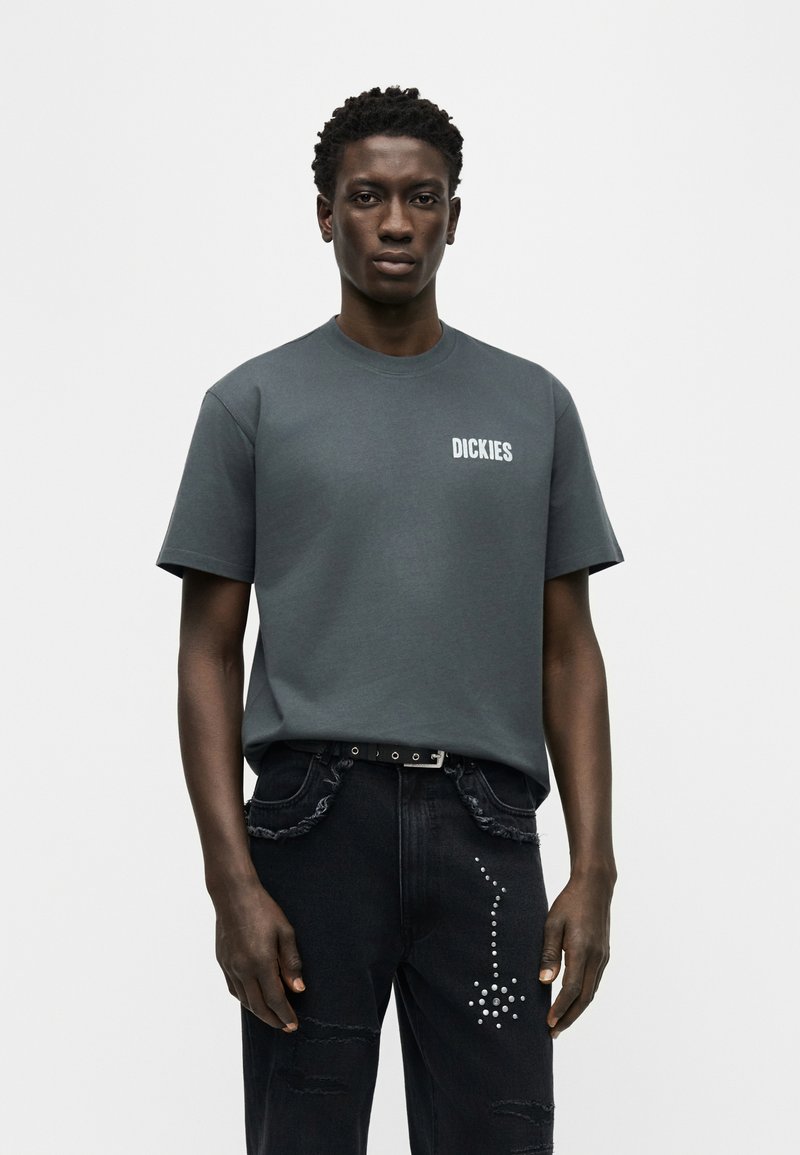 Hombre joven vistiendo una camiseta Dickies gris oscuro y jeans negros con un diseño de flechas tachonadas, posando frente a un fondo claro y liso.