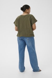 Femme avec les cheveux en chignon, vêtue d'un haut à manches courtes vert olive et d'un jean large bleu, debout dos à un fond blanc.