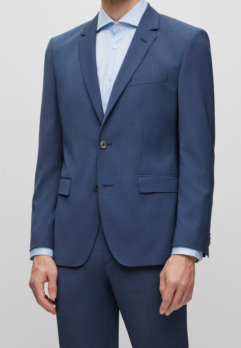 Homme portant un costume bleu ajusté avec une chemise bleu clair, debout devant un fond uni gris clair.