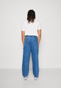 Loose-fit blue denim cargo pants with multiple pockets, a light wash, and a straight leg design. Worn with a plain white t-shirt.