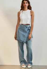 Bijela bezrukavna crop majica, denim suknja na preklop svijetloplave boje, usklađene široke traperice i bijele cipele s crnim točkicama.