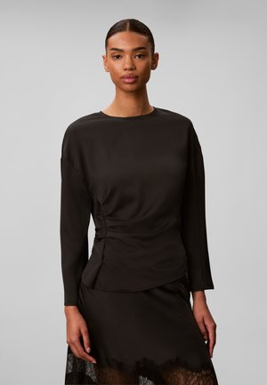 Femme aux cheveux tirés en arrière porte un haut noir à manches longues avec un détail froncé et une jupe noire avec un bord en dentelle.