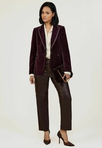 Burgundi bársony blézer selyem hajtókával, krémszínű blúz, barna bőrnadrág és fekete, díszített magassarkú cipő, egy szőrrel díszített táskával a kezében.