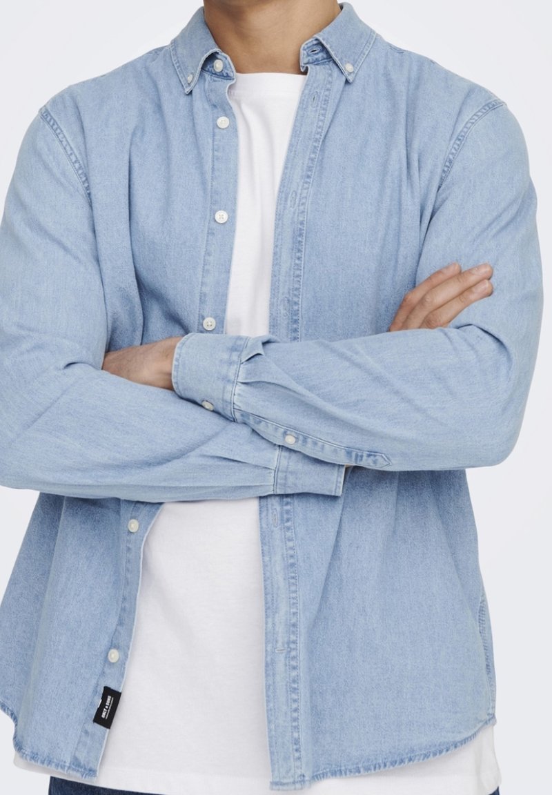 Camicia in denim azzurro chiaro con bottoni, dalla vestibilità rilassata, maniche lunghe e bottoni bianchi, indossata sopra una maglietta bianca. Etichetta visibile di lato.